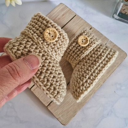Pair of beige knitted booties with buttons on a wooden block.