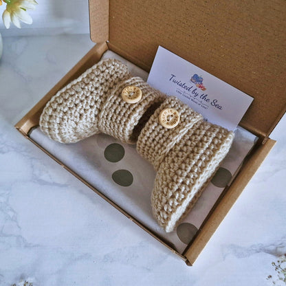 Knitted baby booties with button details in a branded box on a marble surface
