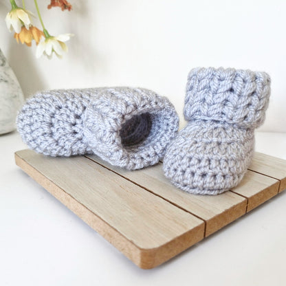 Pair of gray crochet baby booties on a wooden surface with flowers in the background.