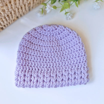 Purple crocheted beanie on a white surface with a woven basket and greenery in the background