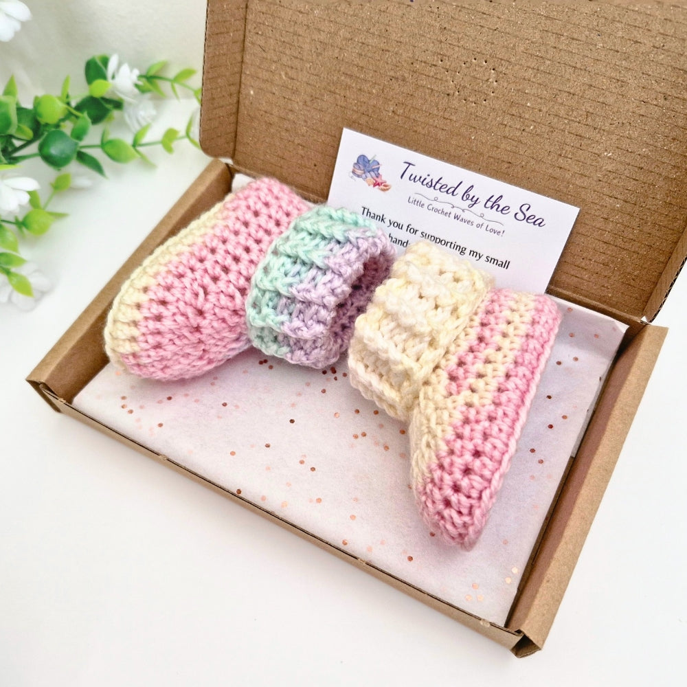 A pair of crocheted booties arranged inside an open brown cardboard gift box with a branded card reading 'Twisted by the Sea.' The booties are pink, mint green, lavender, cream, and pink striped, and the box is photographed on a clean white surface with a small decorative plant in the background.