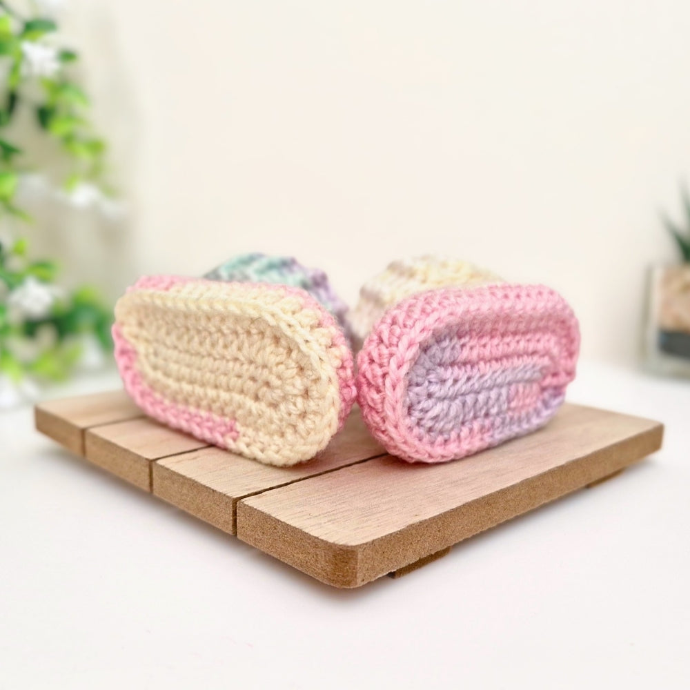 A pair of handmade crocheted baby booties in pastel colors, displayed on a small wooden block against a soft, blurred background.