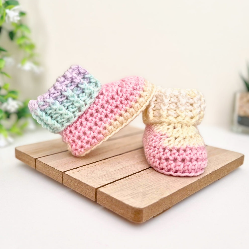 A pair of pastel crocheted baby booties arranged on a small wooden tray against a soft cream background with blurred greenery.