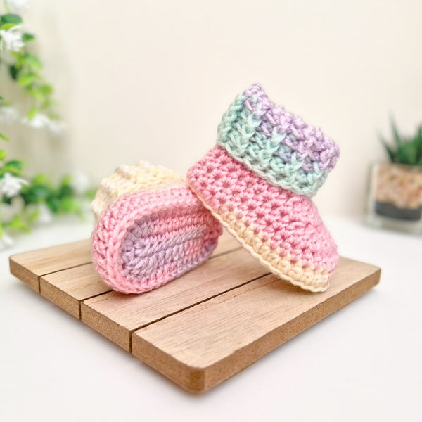 A pair of handmade crocheted baby booties in pastel colors, resting on a small wooden block. The booties feature stripes of pink, light purple, mint green, and cream. Yarn: King Cole Beaches and Cream.