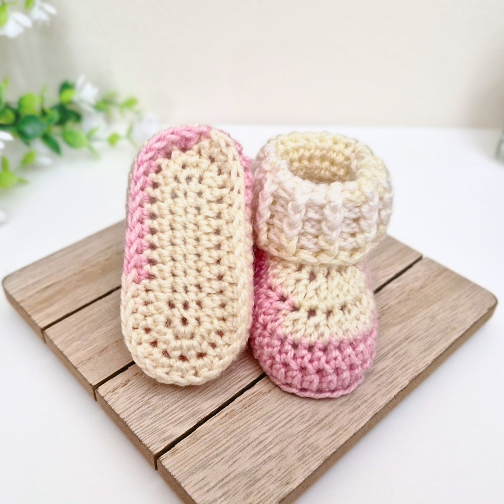 A pair of handmade crocheted baby booties in cream and pink, displayed on a wooden board against a clean white background.