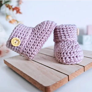Handmade lavender crochet baby booties with wooden button reading Born in 2026 on wooden board