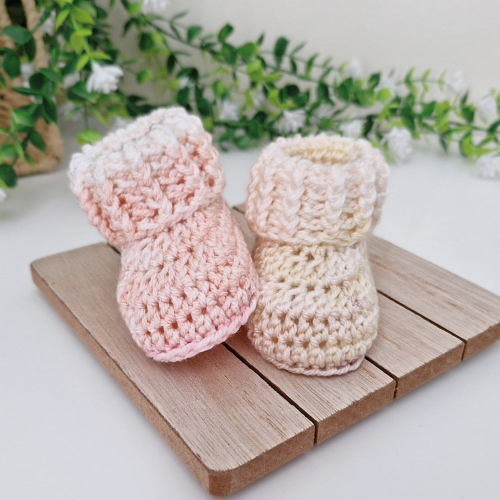 Two crocheted baby booties in soft pastel colors, one light pink and one cream, displayed on a small wooden board with green foliage in the background.