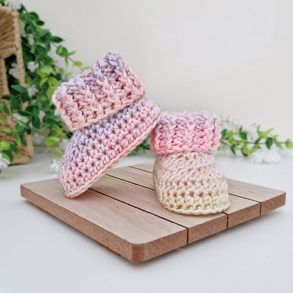 Two handmade crocheted baby booties displayed on a small wooden stand. One bootie is pink and lavender gradient, and the other is cream and pink gradient.