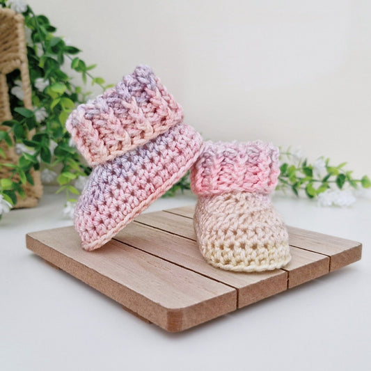 Two handmade crocheted baby booties displayed on a small wooden stand. One bootie is pink and lavender gradient, and the other is cream and pink gradient.