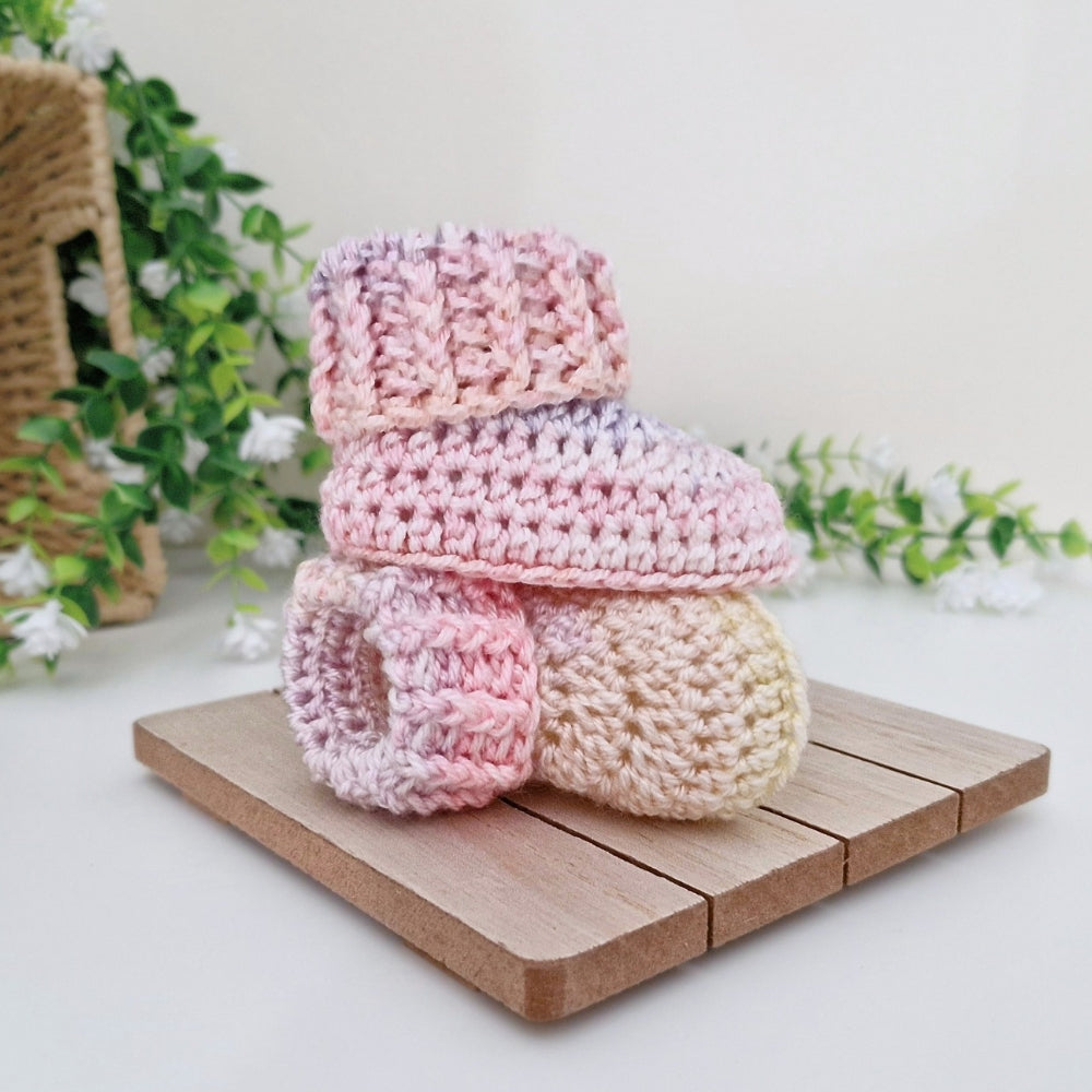 Two pastel crocheted baby booties stacked on a small wooden tray, with a woven basket and greenery in the softly lit background.