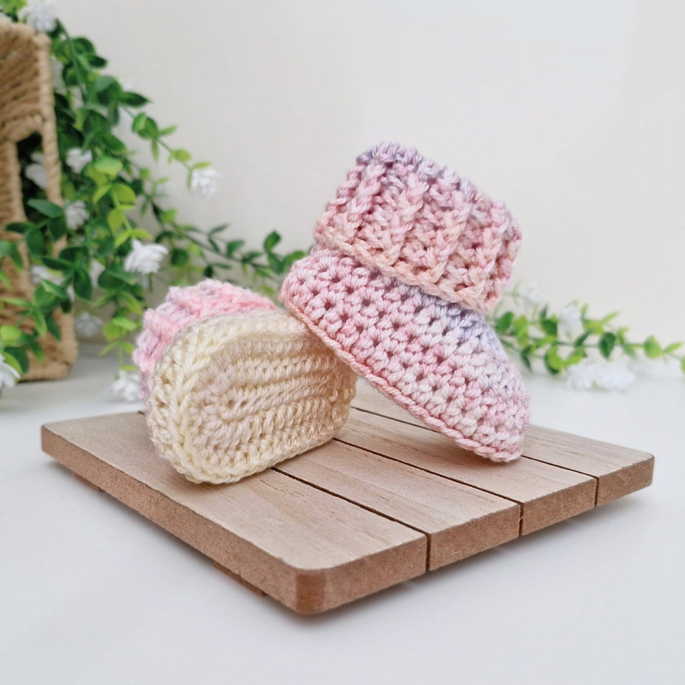 Two crocheted baby booties in pastel variegated colors, one cream and one pink-lavender, displayed on a small wooden block with green foliage in the background.