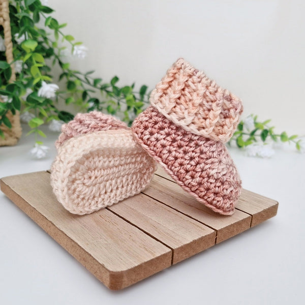 Two handmade crocheted baby booties in soft pastel shades of pink, peach, and cream, displayed on a small wooden block with a soft-focus background of green leaves and white flowers.