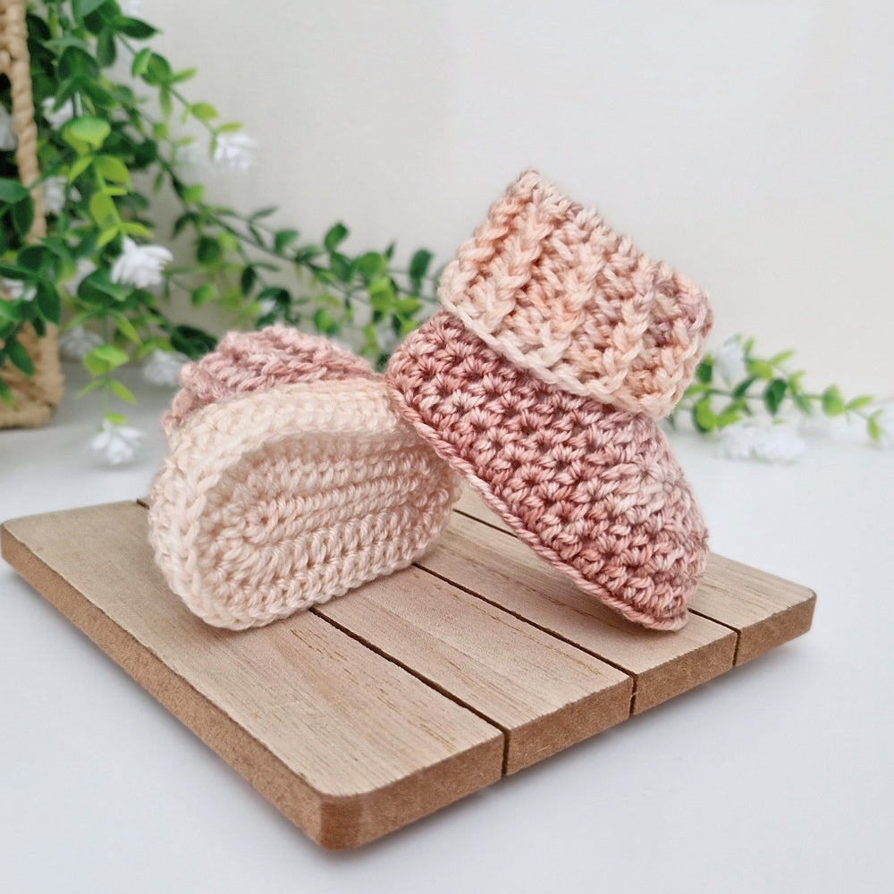 Two handmade crocheted baby booties in soft pastel shades of pink, peach, and cream, displayed on a small wooden block with a soft-focus background of green leaves and white flowers.