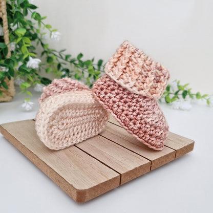 Two handmade crocheted baby booties in soft pastel shades of pink, peach, and cream, displayed on a small wooden block with a soft-focus background of green leaves and white flowers.