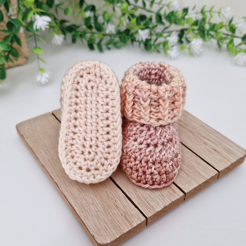 A pair of handmade crocheted baby booties in soft pastel shades, one in light peach-beige and the other in dusty rose, displayed on a small wooden block with green foliage in the background.