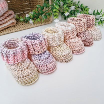 A set of handmade crocheted baby booties in soft pastel colors, including pink, lavender, cream, and peach, arranged on a white surface with a wicker basket and green foliage in the background.