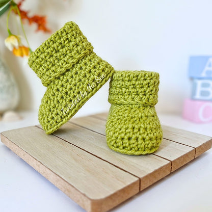 Pair of green crocheted baby booties on a wooden surface with a blurred background.
