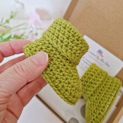 Hand holding a green knitted baby bootie with another bootie in the background.