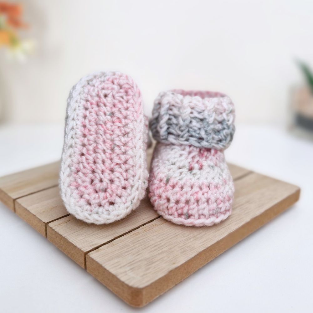 A pair of handmade crocheted baby booties in pink, white, and gray yarn, displayed on a small wooden block against a soft-focus white background.