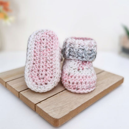 A pair of handmade crocheted baby booties in pink, white, and gray yarn, displayed on a small wooden block against a soft-focus white background.