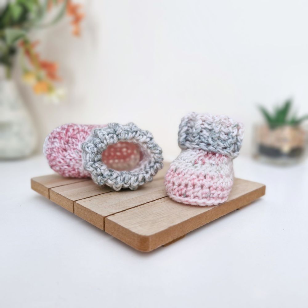 handmade crocheted baby booties in pink and gray yarn, displayed on a small wooden block with a soft-focus background.