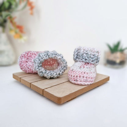 handmade crocheted baby booties in pink and gray yarn, displayed on a small wooden block with a soft-focus background.