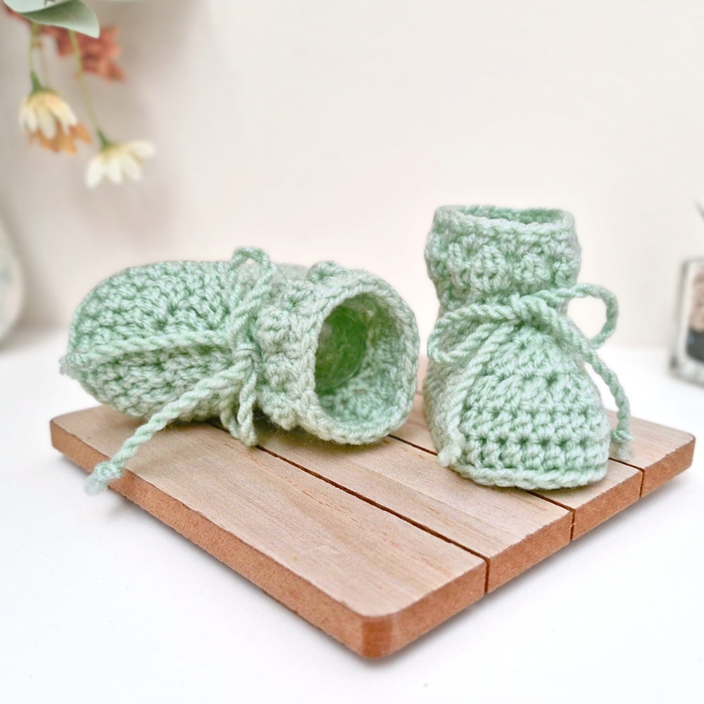 Pair of green knitted baby booties on a wooden board with a blurred floral background