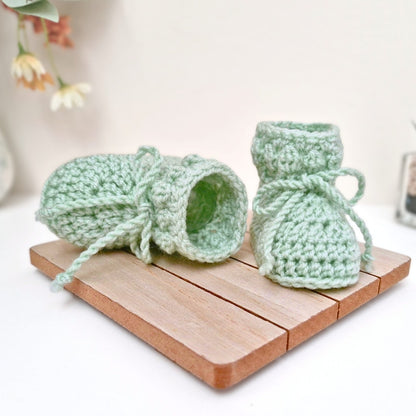 Pair of green knitted baby booties on a wooden board with a blurred floral background