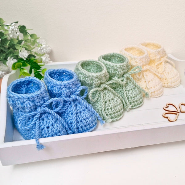 Set of crocheted baby booties in blue, green, and beige on a white surface with a plant in the background.