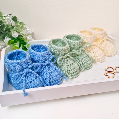 Set of crocheted baby booties in blue, green, and beige on a white surface with a plant in the background.