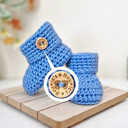 Blue crochet baby booties with a 'It's a Boy' button on a wooden surface.