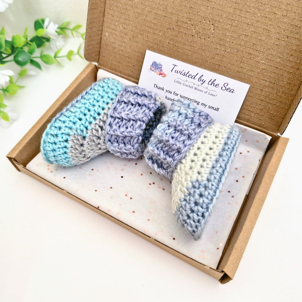 A pair of crocheted baby booties in pastel blue, lavender, and cream are presented in a brown cardboard gift box with a branded card that reads 'Twisted by the Sea'.