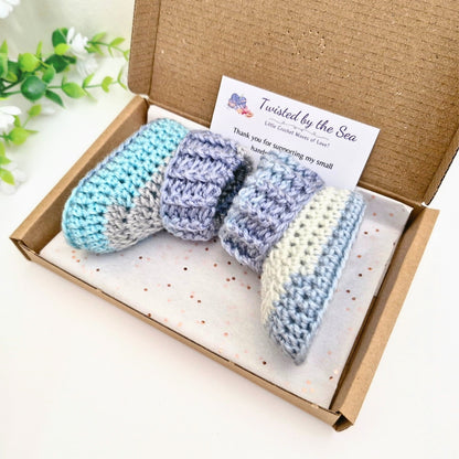 A pair of crocheted baby booties in pastel blue, lavender, and cream are presented in a brown cardboard gift box with a branded card that reads 'Twisted by the Sea'.