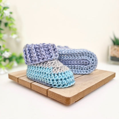 A pair of handmade crocheted baby booties in pastel blue, light gray, and lavender purple, displayed on a small wooden block against a soft, warm background.