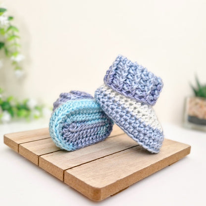 Two crocheted baby booties in pastel blue, lavender, and white, displayed on a small wooden board against a softly blurred light background with decorative plants.