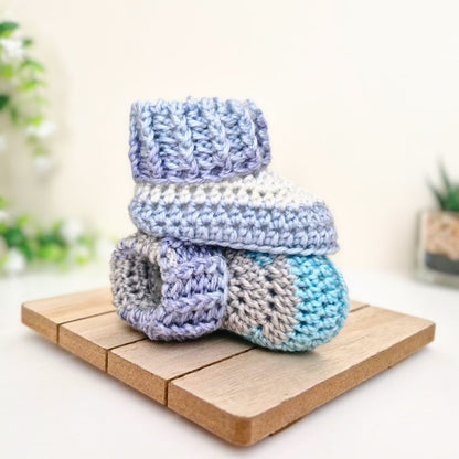 A pair of handmade crocheted baby booties stacked on a small wooden board, featuring pastel stripes of lavender, white, light blue, and turquoise. Yarn: King Cole Palm Beach.