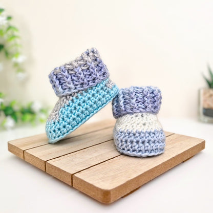 A pair of handmade crocheted baby booties in pastel blue, lavender, and white, displayed on a small wooden block against a soft, warm background.