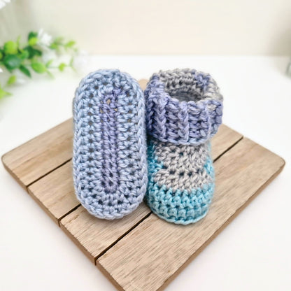 A pair of handmade crocheted baby booties in pastel shades of lavender, light blue, and gray, displayed on a wooden block.