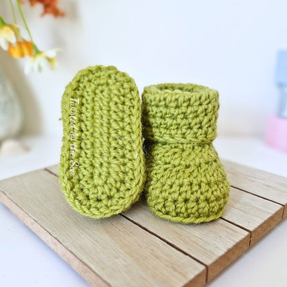 Pair of green knitted baby booties on a wooden surface with a blurred background.