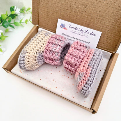 A pair of handmade crocheted baby booties in pastel pink, lavender, and cream, presented inside a brown cardboard gift box with a branded card. The background is a clean white surface with a decorative green plant in the corner.