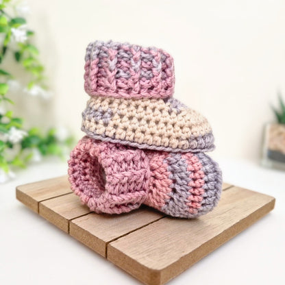 A pair of handmade crocheted baby booties stacked on a small wooden block, featuring a color-blocked pattern of pink, beige, and light purple, with a soft-focus background showing green plants.