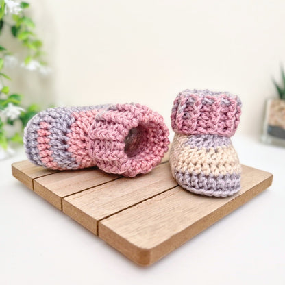 A pair of handmade crocheted baby booties in pink, lavender, and cream, displayed on a small wooden block with a soft-focus background.