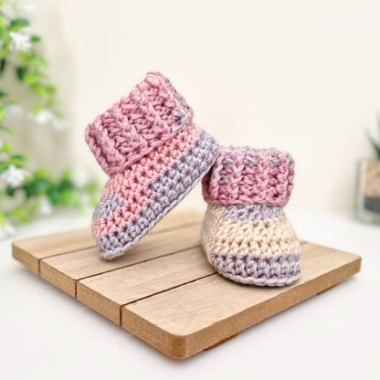 A pair of handmade crocheted baby booties in pink, lavender, and cream, displayed on a small wooden block against a soft, blurred background. Yarn is King Cole Pebble Beach
