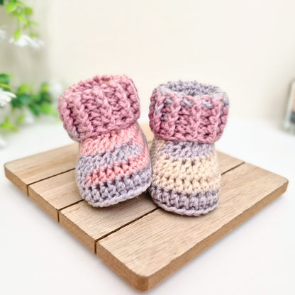 A pair of handmade crocheted baby booties in pastel pink, lavender, and cream, displayed on a small wooden block.
