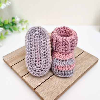 Two crocheted baby booties displayed on a small wooden board against a clean white background with soft green leaves in the corner. One bootie is light lavender-gray, and the other is pink and lavender-gray striped.