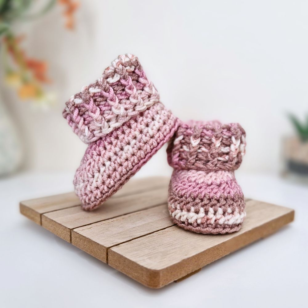A pair of hand-crocheted baby booties in a variegated pink and cream yarn, displayed on a small wooden block against a soft-focus white background.