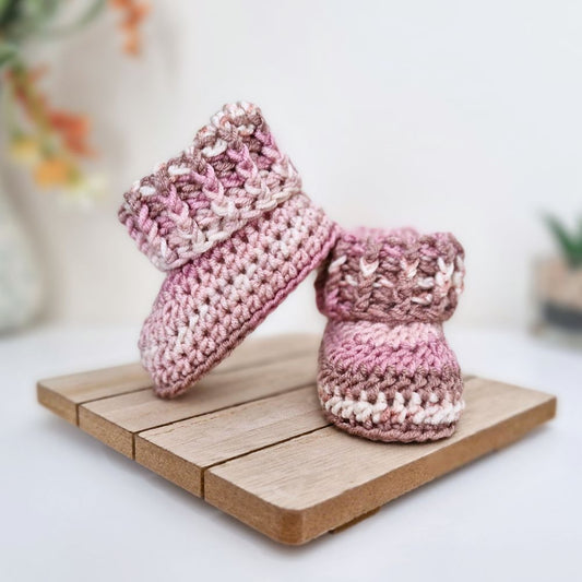 A pair of hand-crocheted baby booties in a variegated pink and cream yarn, displayed on a small wooden block against a soft-focus white background.