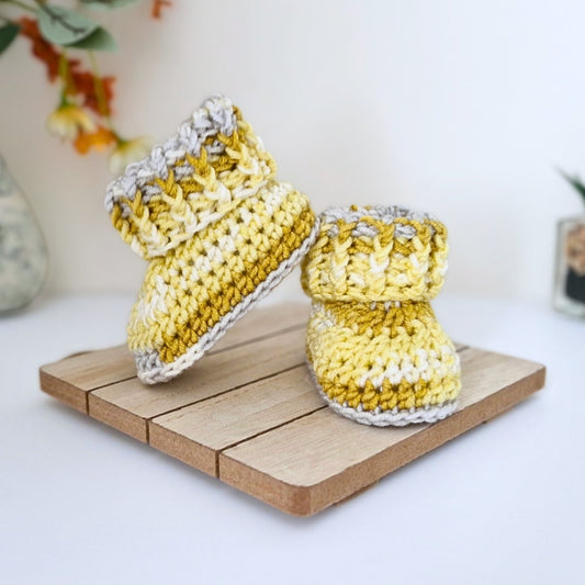 A pair of handmade crocheted baby booties in yellow, white, and gray yarn, displayed on a small wooden block against a clean white background.