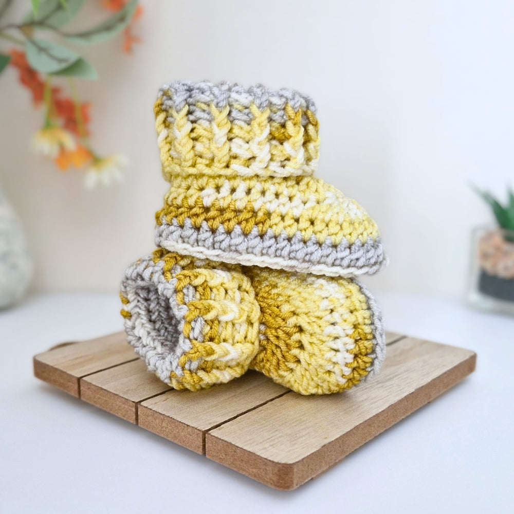 handmade crocheted baby booties in yellow and gray, stacked on a small wooden block against a bright, softly blurred background.