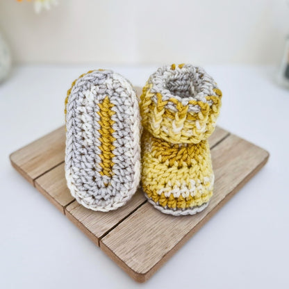 A pair of handmade crocheted baby booties in yellow and white, displayed on a small wooden block against a clean white background.